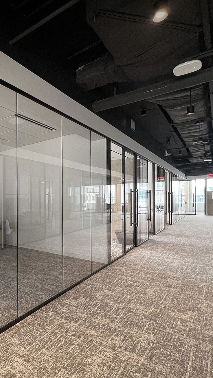 Modern office corridor with glass-walled conference rooms, black ceiling fixtures, and carpeted floor