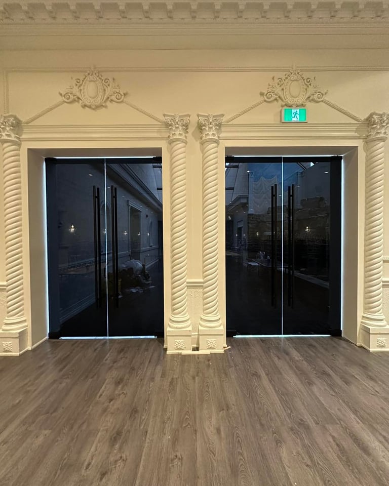 Two elegant glass doors with cream ornamental columns and exit sign above on light wood flooring