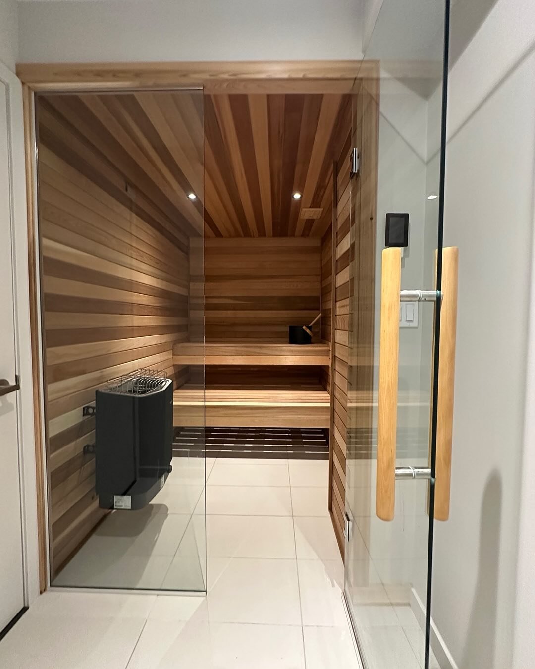 Modern sauna with wood paneling, benches, and heating stove inside glass doors in a bathroom